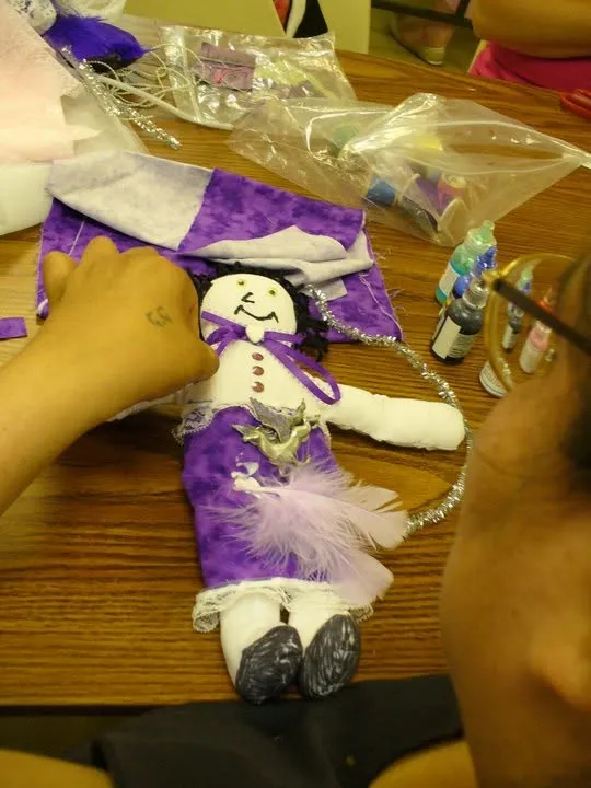 A maker adding ribbon and fabric to a cloth doll at a table of craft materials