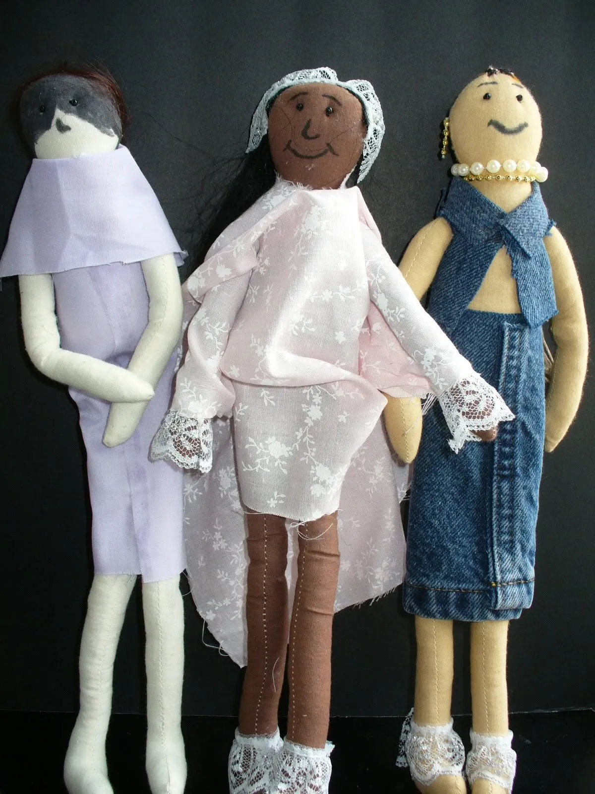 Three handmade cloth dolls with individual expressions, dressed in lilac fabric, pink lace, and blue denim